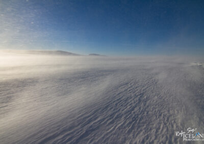 Snow-covered landscape with strong winds blowing snow across a flat terrain under a clear blue sky.