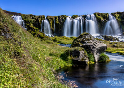 Waterfalls cascading over a rocky ledge with moss-covered boulders, surrounded by lush greenery under a clear blue sky.