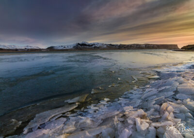 The tranquil shoreline of a lake in Iceland, adorned with ice and snow, captures the essence of a frosty landscape.