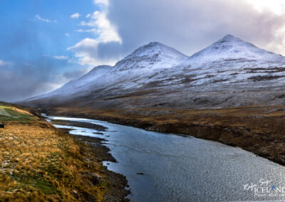 A serene river flows through a grassy field, framed by snow-capped mountains in the stunning landscape of Iceland.