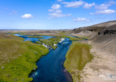 A picturesque river flows through a verdant valley, with towering mountains rising in the background, showcasing Iceland's beauty and the black volcanic sand