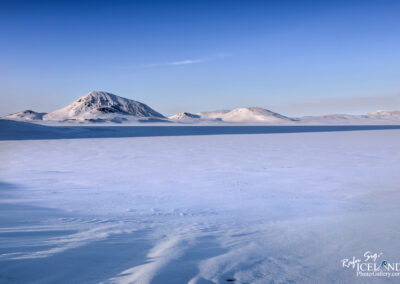 An idyllic winter landscape in Iceland, with snow-covered terrain and towering mountains visible in the distance beneath a clear blue sky