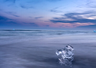 An iceberg sits on a black sandy beach, bathed in the golden light of a sunset, creating a serene coastal scene.