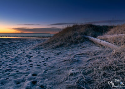 A picturesque black sand beach scene at sunset, highlighting a log on the sand and rolling dunes under a colorful sky.