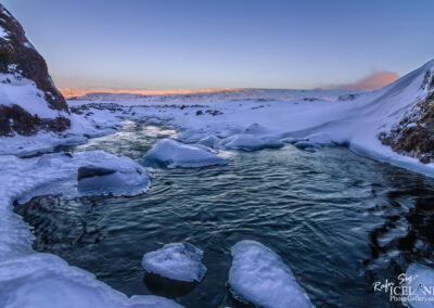 A serene river meanders through a tranquil snowy landscape and rocks, reflecting the beauty of winter's embrace.