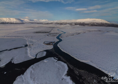 A serene river meanders through a tranquil snowy field, reflecting the calmness of winter's embrace.