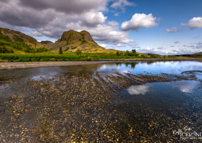 A small, calm water body nestled among rocks, mountains and green grass, creating a picturesque outdoor scene with little water in the foreground.