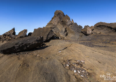 A rocky landscape featuring large, jagged boulders and varied textures on the ground. The rocks are dark and rugged, set against a clear blue sky. The foreground shows a mix of rock sizes, with some displaying white patches, suggesting the presence of minerals or biological growth. The background features further rocky formations that rise prominently.
