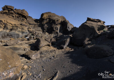 A rugged rocky landscape with large, uneven rocks and boulders of various sizes scattered across the ground. The rocks are a mix of brown and gray tones, with some showing signs of weathering and moss growth. In the background, taller rock formations rise upwards against a clear blue sky, creating a dramatic natural scene. The foreground features a sandy and pebbly surface, further emphasizing the rugged terrain.