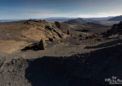 A vast, rugged landscape featuring dark volcanic rock formations and a sloping terrain. In the foreground, angular rocks are scattered across a rocky surface, leading towards a distant horizon. The background shows rolling hills and mountains under a clear blue sky, with a hint of water visible in the distance. The lighting creates soft shadows, highlighting the textures of the rocky ground.