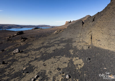 A rocky, black volcanic landscape stretches out under a clear blue sky. In the background, a serene lake reflects the surroundings, while rough terrain and scattered stones dominate the foreground. The slopes of the land rise gently, revealing varied textures and colors, with dark, textured surfaces contrasting against lighter patches. The scene conveys a sense of rugged natural beauty.