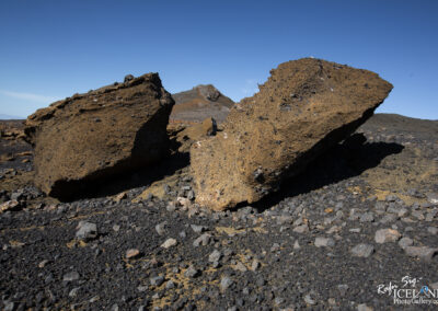 A barren, rocky landscape featuring two large volcanic rocks in the foreground. The rocks are textured and rugged, with one displaying a rough, dark surface and the other appearing more yellowish-brown. The ground is covered in small pebbles and volcanic debris, with a clear blue sky above and a distant rocky formation in the background.