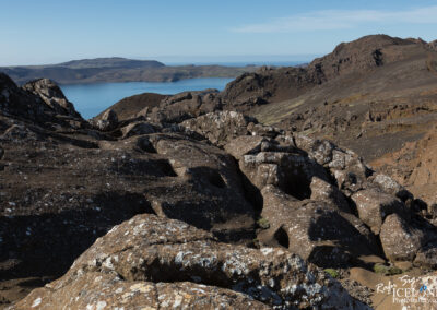 A panoramic view of rugged, rocky terrain with various sizes of brown and gray rocks in the foreground. In the background, a calm lake reflects the blue sky, surrounded by distant hills and mountains. The landscape appears arid and features some patches of green vegetation amidst the rocky surfaces.