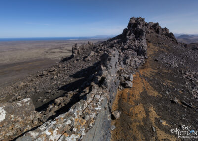 A rocky landscape view from a high vantage point, showcasing jagged cliffs and boulders in the foreground. The terrain is predominantly barren and volcanic, featuring dark rocks and patches of orange-brown earth. In the distance, a vast expanse of flat land stretches towards the horizon, meeting the blue sea under a clear sky. The distant coastline is visible, with faint outlines of land and mountains further away.