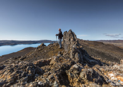 A person stands on a rocky ridge overlooking a tranquil blue lake and distant rolling hills. The landscape is rugged, with jagged rocks in the foreground and a clear blue sky above. The individual holds a walking stick, dressed casually and appearing to enjoy the scenic view.