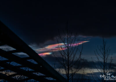 A dramatic twilight sky featuring dark clouds with a streak of colorful, iridescent clouds displaying shades of pink and blue. Silhouette of bare trees is visible in the foreground, along with part of a structured framework or lattice. The overall scene conveys a serene and captivating atmosphere.