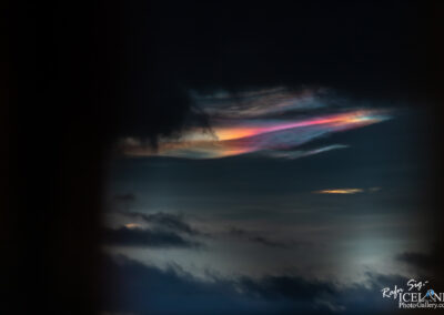 A dramatic sky featuring dark clouds with a vibrant, iridescent band of color near the horizon. The hues transition from deep shades to bright pink and orange, creating a striking contrast against the darker backdrop of the clouds. The overall scene evokes a sense of awe and wonder, capturing the beauty of natural light phenomena.
