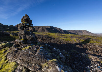 A stone cairn stands prominently in the foreground, composed of unevenly stacked dark rocks, some adorned with patches of green moss. The background features expansive landscapes with rolling hills and rugged terrain under a clear blue sky, while the horizon showcases a low mountain range. The scene conveys a sense of tranquility and natural beauty.