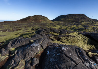 A rugged landscape characterized by dark, rocky terrain interspersed with patches of vibrant green moss. In the background, two volcanic hills rise against a clear blue sky, showcasing their steep, textured slopes. The foreground features cracked lava formations, adding depth and contrast to the scene. The overall atmosphere is one of natural beauty and geological intrigue.