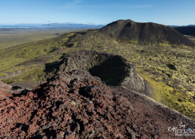 A panoramic view of a volcanic landscape features rugged, black lava rock formations in the foreground, along with a large volcanic crater. In the background, rolling hills and mountains are visible under a clear blue sky, with patches of green moss covering the land. The scene conveys a sense of vastness and the natural beauty of a volcanic region.