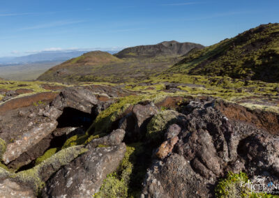 A scenic landscape featuring rugged volcanic terrain with dark, rocky surfaces partially covered in vibrant green moss. In the background, rolling hills and distant mountains stretch under a clear blue sky, while patches of clouds can be seen above the horizon. The foreground includes a noticeable crevice in the ground, adding depth to the natural features of the environment.