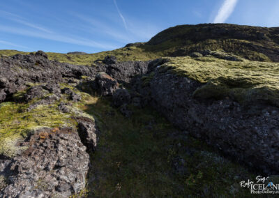A rocky landscape covered in bright green moss, with rugged dark volcanic rocks scattered throughout. In the background, a gentle hill rises under a clear blue sky, with wispy clouds streaking across the horizon. The foreground features patches of grass peeking through the moss, enhancing the natural beauty of the scene.