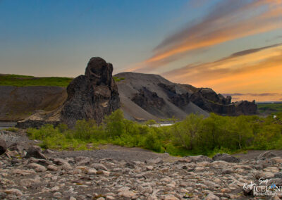 A dramatic landscape featuring rugged rock formations and rolling hills, with a foreground of scattered rocks and boulders. The evening sky showcases soft hues of orange and blue, suggesting twilight. Lush greenery intersperses the rocky terrain, hinting at a river flowing nearby. The scene captures the raw beauty of nature in a remote area, likely in Iceland.