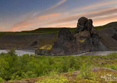 A rugged landscape featuring a prominent rock formation alongside a winding river. The terrain is dotted with lush green foliage and distant hills, under a colorful sky with soft sunset hues. The rocky outcrop displays textured surfaces and moss growth, contrasting with the smoother riverbank.