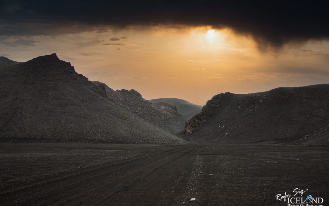 A barren landscape featuring dark, rocky hills under an overcast sky. The sun is partially obscured by clouds, casting a warm golden light that contrasts with the darker terrain. The ground is uneven and marked with tire tracks, leading into a narrow valley formed by the surrounding hills.