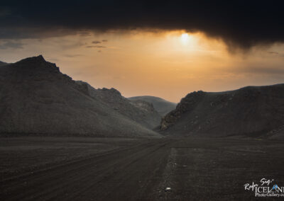 A barren landscape featuring dark, rocky hills under an overcast sky. The sun is partially obscured by clouds, casting a warm golden light that contrasts with the darker terrain. The ground is uneven and marked with tire tracks, leading into a narrow valley formed by the surrounding hills.