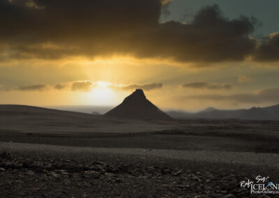 A barren landscape at sunset, featuring a distinctive conical hill silhouetted against the vibrant hues of the sky. The foreground is rocky and desolate, and the background showcases rolling mountains fading into the distance under a mix of clouds and light. The sky transitions from blue to golden yellow as the sun sets on the horizon.
