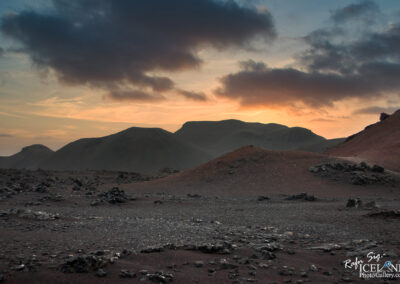 A dramatic landscape featuring a barren area with reddish-brown earth and scattered black volcanic rocks. In the background, there are several smooth, rolling mountains under a moody sky filled with clouds. The scene captures the soft, warm glow of a sunset, highlighting the contours of the terrain.