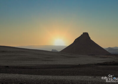 A sunrise illuminates a landscape featuring a prominent, pyramid-shaped hill surrounded by a barren, rocky terrain. The sky transitions from pale blue to soft yellow and orange hues, casting a serene atmosphere over the scene.