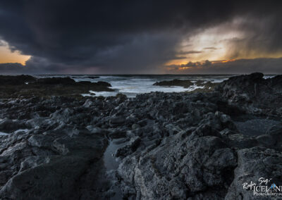 A dramatic coastal scene at twilight, featuring rugged black volcanic rocks in the foreground, with turbulent ocean waves crashing against them. The sky is filled with dark, ominous clouds and hints of orange and yellow from the setting sun breaking through, creating a contrast between the stormy atmosphere and the warmth of the sunset. The overall mood is both powerful and serene, showcasing the beauty and intensity of nature.