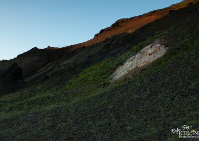 A sloped landscape with rugged terrain featuring dark volcanic rock and patches of green grass. At the center, there is a lighter section of exposed earth, contrasting with the surrounding dark soil. The background shows a rocky ridge against a clear blue sky, illuminated by soft sunlight, suggesting early morning or late afternoon light.