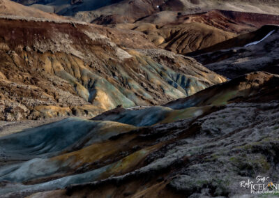 A rugged, multicolored volcanic landscape featuring a series of rolling hills and valleys. The terrain exhibits various shades of brown, green, and orange, creating a striking visual contrast. Sparse vegetation is visible in some areas, while others appear more barren. The lighting suggests a scene captured during the late afternoon or early evening, enhancing the textures and colors of the geological formations.