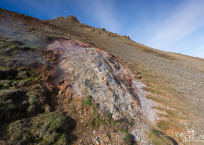 A rugged landscape featuring a colorful, mineral-stained rocky outcrop with steam rising from it, set against a backdrop of a sloping, rocky terrain under a clear blue sky. The foreground has patches of green grass interspersed with exposed earth and gravel. The overall scene conveys a sense of natural geothermal activity.