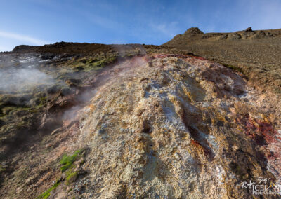 A colorful, mineral-rich landscape with a rocky hillside partially covered in green moss. Steam is rising from the ground, indicating geothermal activity. The sky above is clear with a few wispy clouds. The terrain displays various hues of yellow, orange, and red, showcasing the unique geological features of the area.