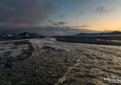 A serene landscape featuring a winding river flowing through a rocky terrain under a cloudy sky. In the background, mountains with patches of snow are visible, and the horizon glows with the soft light of sunset, casting a calming ambiance over the scene. The water appears slightly murky, reflecting the colors of the sky.