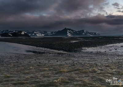 A rugged landscape with large, dark mountains in the background, some capped with snow. The foreground features a flowing river with a mix of calm and slightly turbulent water, reflecting the muted colors of the sky. The atmosphere is dramatic with dark clouds overhead, suggesting a moody or impending weather change. The scene is devoid of vegetation, emphasizing the stark, natural beauty of the terrain.