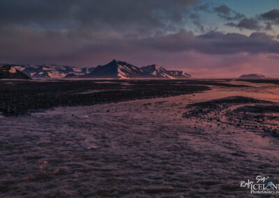 A scenic landscape featuring a river flowing through a rocky terrain, with mountains in the background partially covered in snow. The sky displays a blend of colors from sunset, casting a warm glow over the mountains and water. The scene captures the serene beauty of a remote, natural environment.