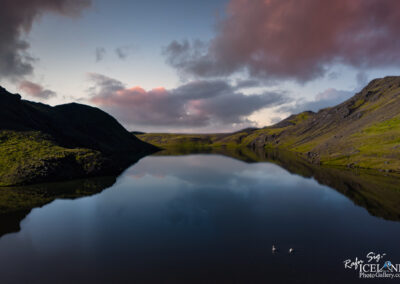 A serene landscape features a calm lake reflecting the surrounding hills and sky. The water's surface is smooth, with gentle ripples created by three swans gliding across. The hills are lush and green, interspersed with patches of rocky terrain, while soft clouds in various shades of gray and pink hover above. The scene evokes a peaceful, natural environment.