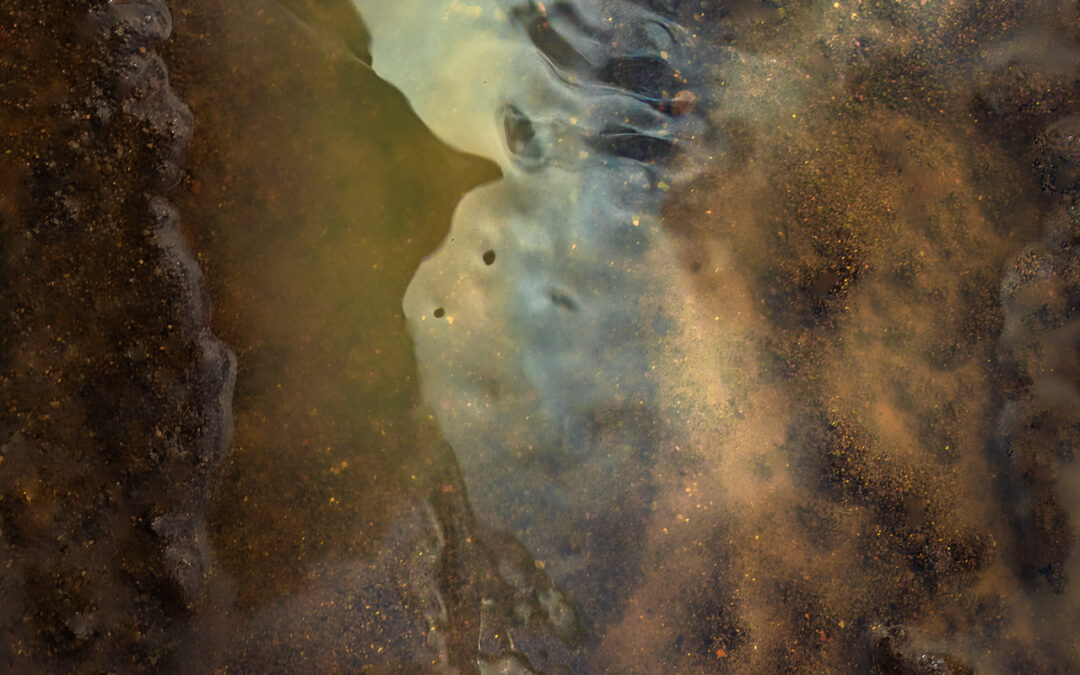 The image features a close-up view of a reflective, muddy surface, where light diffuses through water, creating an abstract blend of earthy tones and soft blues. The texture appears rough with elements of dirt and smooth water, capturing the intricate details of natural surroundings.