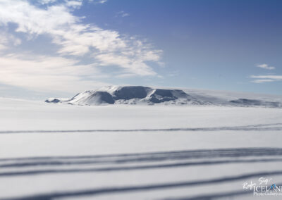 A vast, snow-covered landscape stretches into the distance under a clear blue sky. Gently rolling hills and a prominent mountain are visible, their peaks dusted with snow. Thin clouds float in the sky, creating a serene and tranquil atmosphere. The foreground features subtle shadow lines across the smooth snow, indicating the presence of previous footprints or tracks.