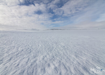 A vast expanse of snow covers the ground, showing subtle patterns and textures. The scene is illuminated by soft, diffuse light under a sky filled with scattered clouds, ranging from white to shades of gray. In the distance, gentle hills rise against the backdrop of a light blue sky. The overall mood is tranquil and serene, emphasizing the quiet beauty of a snowy landscape.
