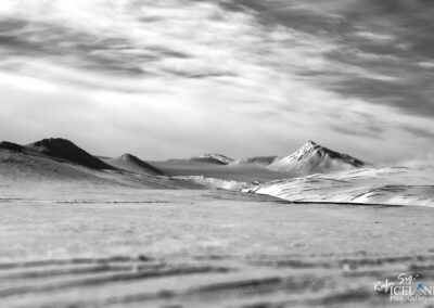 A black and white landscape featuring rolling hills and mountains under an overcast sky. The foreground shows a smooth, snow-covered terrain with gentle ridges and shadows. In the background, several sharp peaks rise, some partially shrouded in mist. The contrast between the bright snow and the darker mountains creates a dramatic scene.
