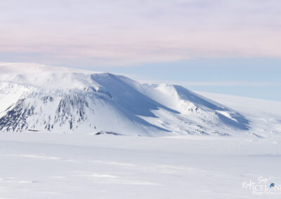 A serene winter landscape featuring snow-covered mountains and a vast expanse of white terrain under a soft pastel sky. The peaks are sharply defined, showcasing undulating ridges and shadows cast by the sunlight, while the foreground highlights the untouched, smooth snowy surface.