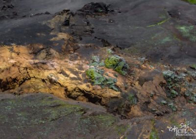 A rugged and colorful landscape featuring a mix of dark volcanic rock, patches of yellow and orange earth, and vibrant green and blue lichen. The terrain shows a variety of textures and colors, indicating geological activity and diverse vegetation. The scene captures the stark beauty of a natural environment shaped by volcanic processes.