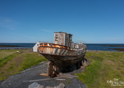 An old, weathered fishing boat sits on a grassy area near the coast. The boat, painted white with rusted patches, is positioned on wooden supports. In the background, a small white building is partially visible, along with a rocky shoreline and the blue sea under a clear sky. The landscape is dotted with yellow wildflowers, and a few scattered rocks can be seen along the shore.