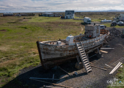 A weathered wooden boat, aged and partially dismantled, is prominently featured in the foreground, resting on a rocky surface with a set of stairs leading up to its deck. In the background, a green landscape stretches out, dotted with a few houses and vehicles. The sky is partly cloudy, suggesting a calm, open atmosphere. The surrounding area appears to be rural, with grassy fields and a distant view of hills or mountains.